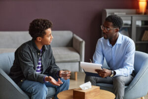 teen boy sits in a chair talking with a therapist about process addiction treatment.